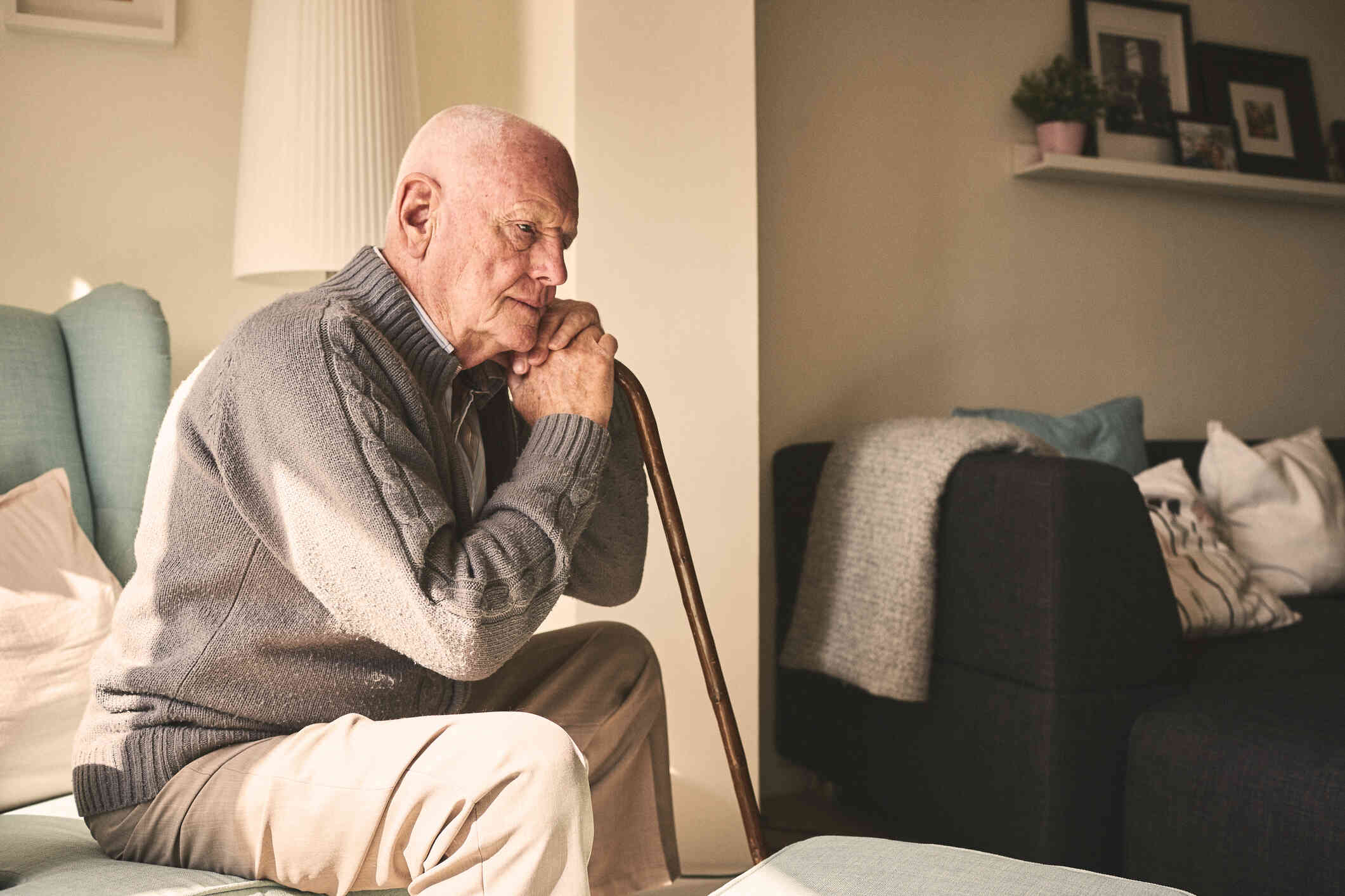 An elderly man in a tan sweater sits on a chair in his home and sadly rests his head against his cane while gazing off with a sad expression.